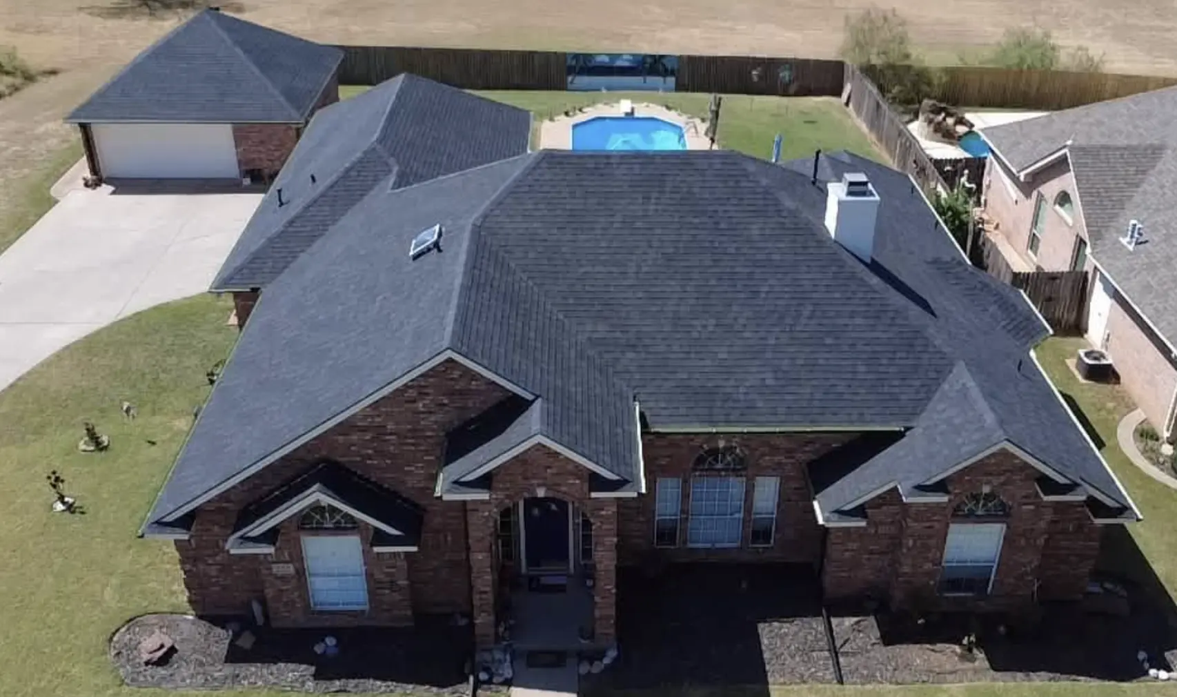 Aerial view of a residential roofing project in Bismarck, North Dakota, showing a completed dark asphalt shingle roof on a brick home with a chimney and pool by Bismarck Exterior Pros.